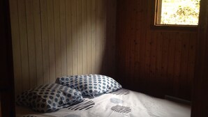 Une petite pièce à l’éclairage tamisé, avec un lit simple, des murs en bois et une fenêtre laissant entrer la lumière naturelle.