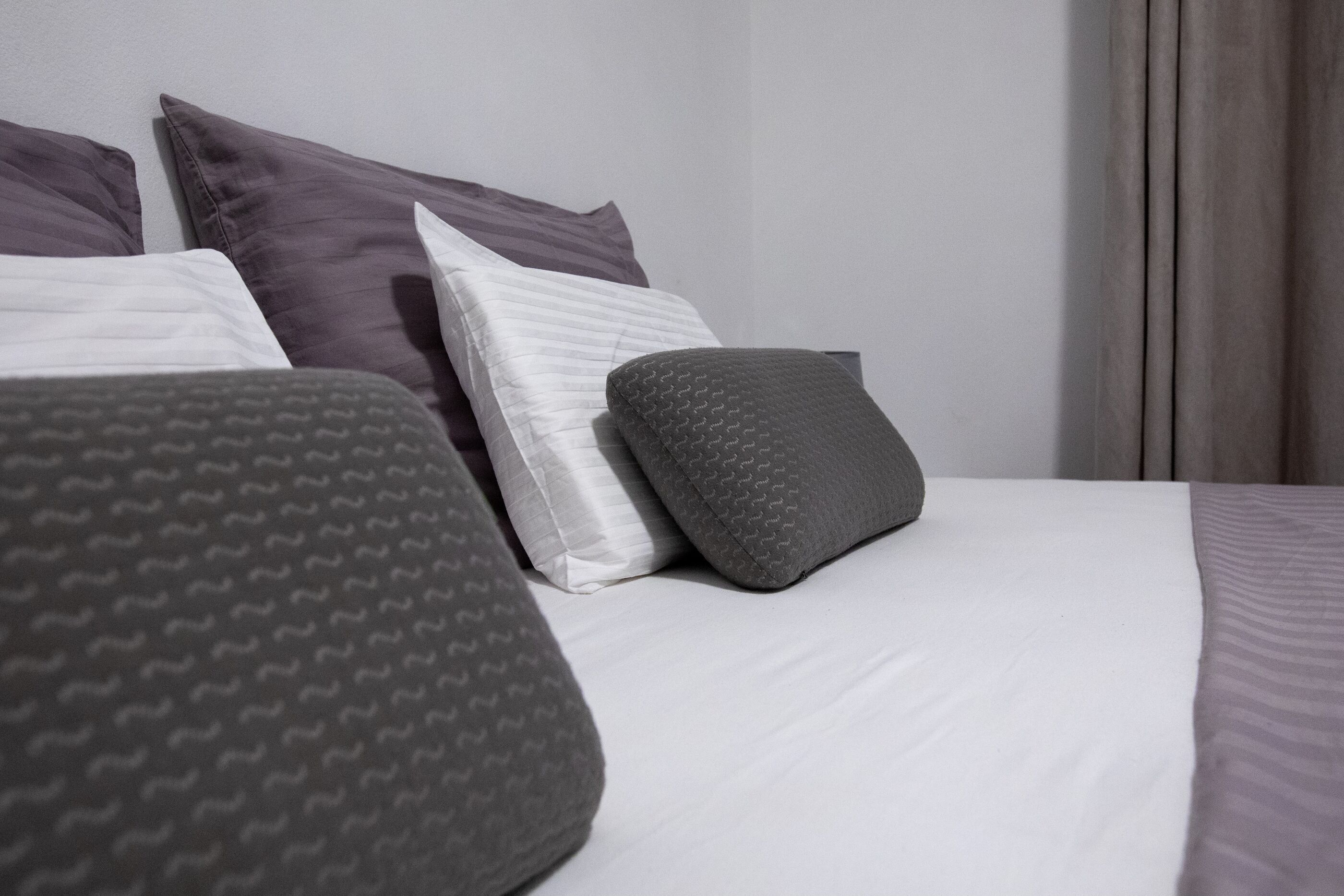 Uma cama bem arrumada com almofadas e um cobertor, cabeceira e janela com cortinas.