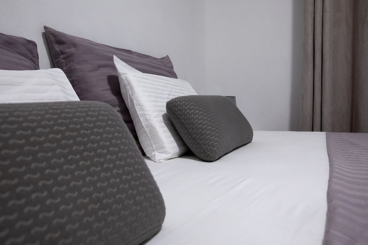 Una cama bien hecha con ropa de cama blanca y gris, incluyendo almohadas y una manta.