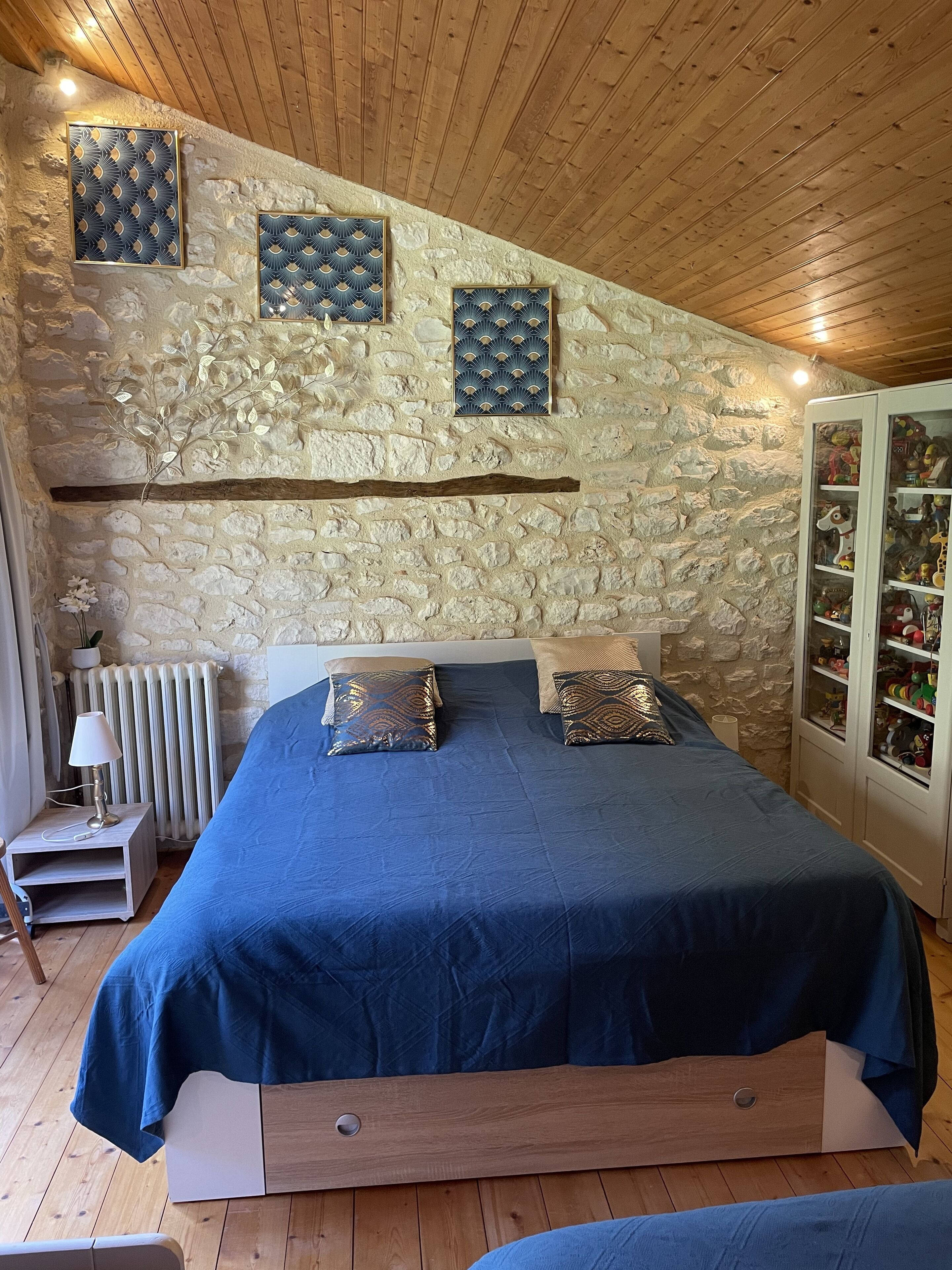 A bedroom with a stone wall, a wooden ceiling, a bed with blue bedding, and a cabinet with toys.