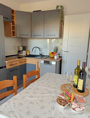 A compact kitchen with modern appliances, a dining table with a floral tablecloth, and a wine bottle on the table.