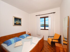 A bedroom with a wooden bed, a small table with a lamp, a chair, a window with a view of the sea, and a painting on the wall.