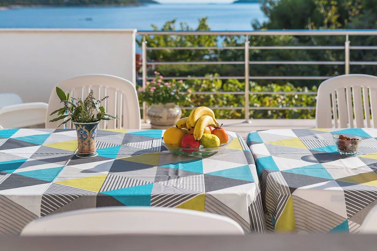 A table with a geometric patterned tablecloth, white chairs, a potted plant, and fruit on the table.