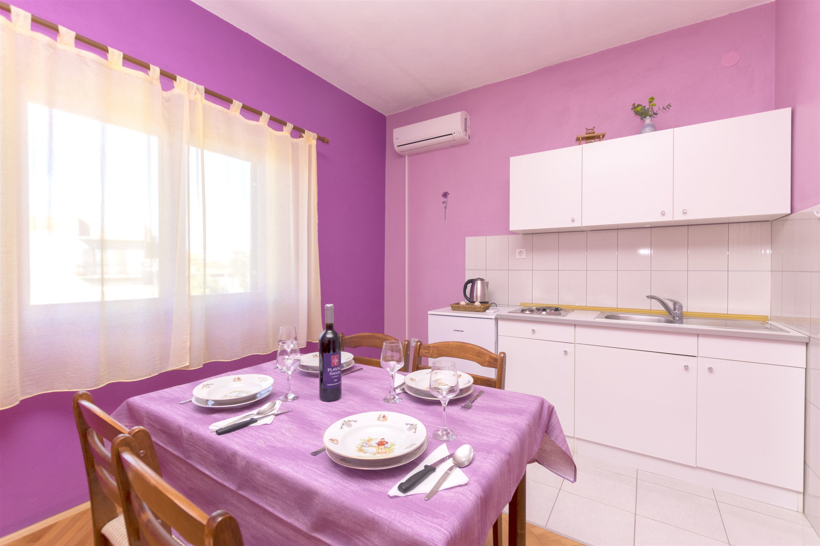 Un coin repas avec une nappe violette, des meubles blancs et une fenêtre ornée de rideaux légers.