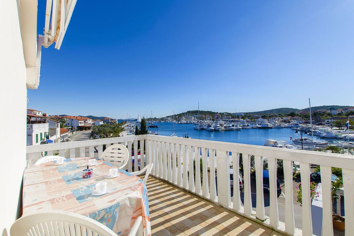Ein Balkon mit Esstisch, der auf eine Marina mit Booten und eine hügelige Landschaft blickt.