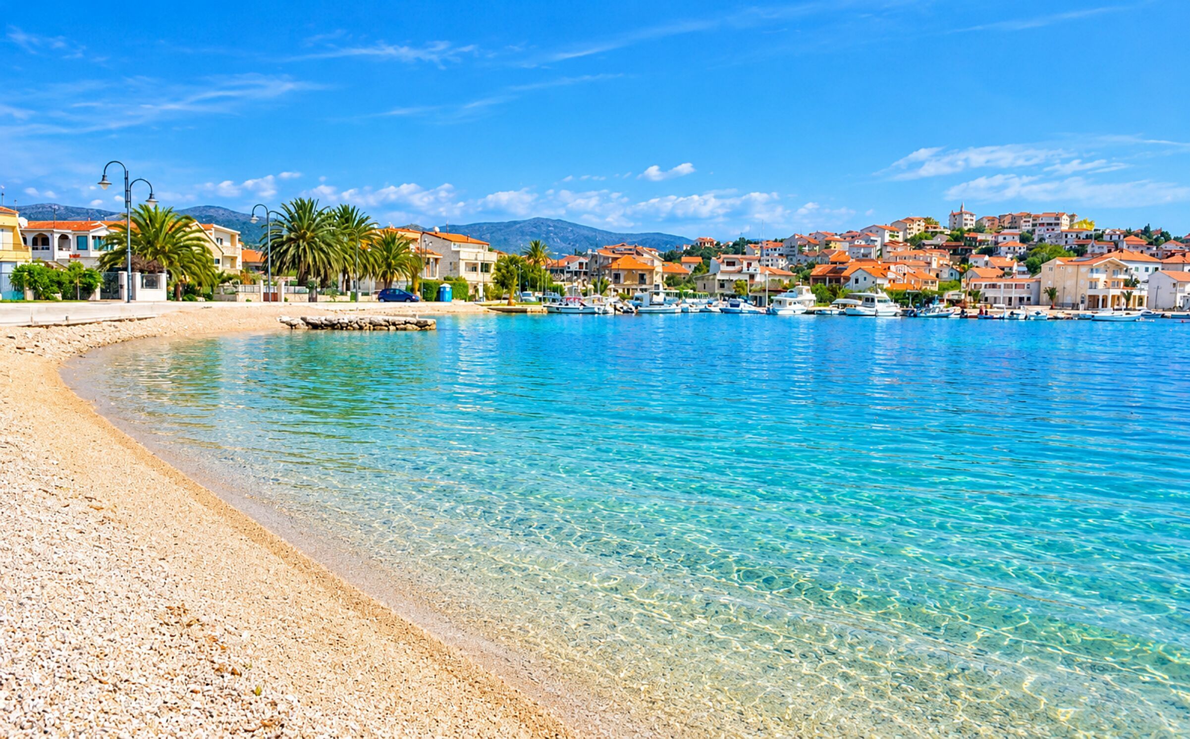 A coastal town with clear blue waters, a pebble beach, and buildings with red roofs.