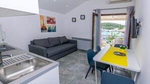 Een knus woongedeelte met een kitchenette, een grijze bank, een eettafel met stoelen en uitzicht op een jachthaven vanaf een balkon.