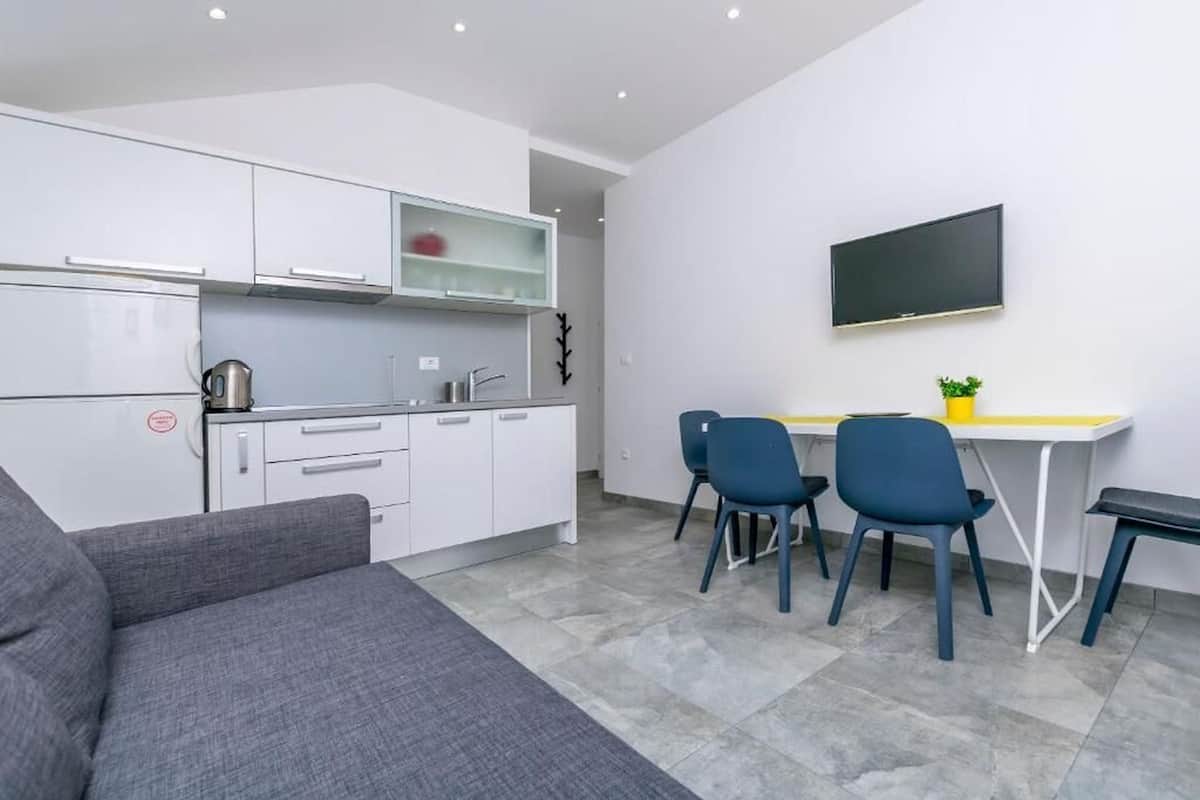 A modern kitchen with white cabinets, a kettle, and a sink. There is a dining area with two blue chairs and a small table. A grey sofa is in the foreground.