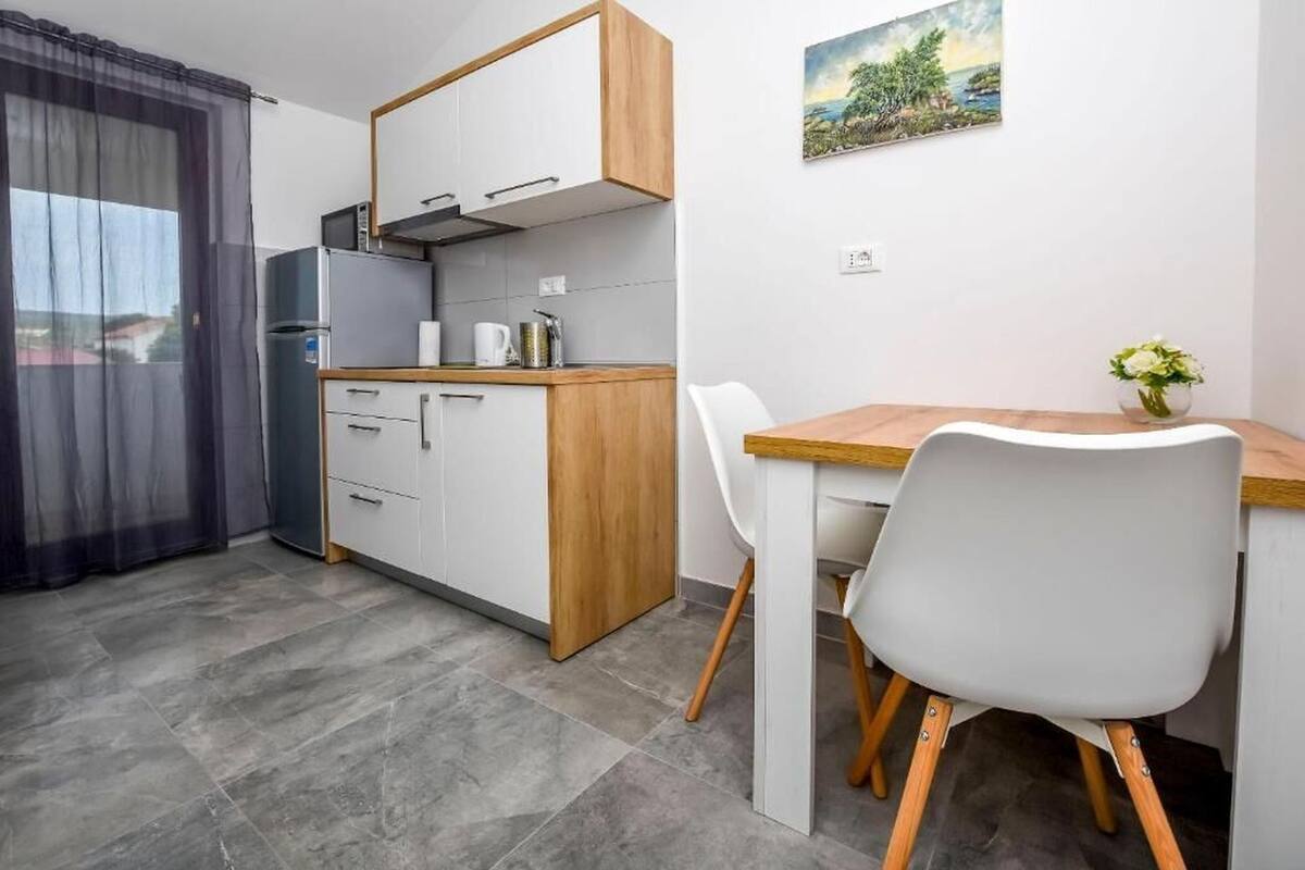 A compact kitchen with a refrigerator, microwave, and wooden cabinets. There's a dining table with chairs and a small vase with flowers.