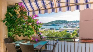 Een balkon met rieten meubilair, een gestreepte luifel en uitzicht op een jachthaven en heuvels.