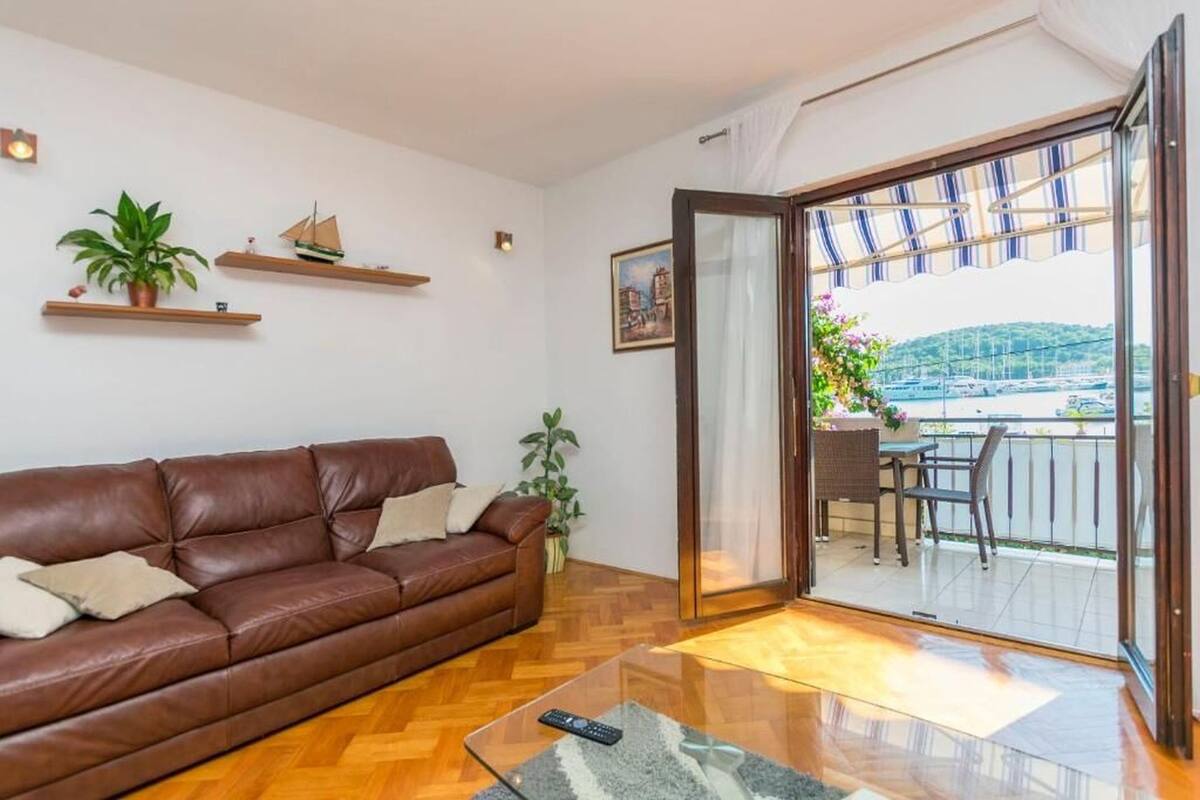 A living room with a brown leather sofa, wooden floor, and a glass coffee table. There is a balcony with a view of the sea and boats.