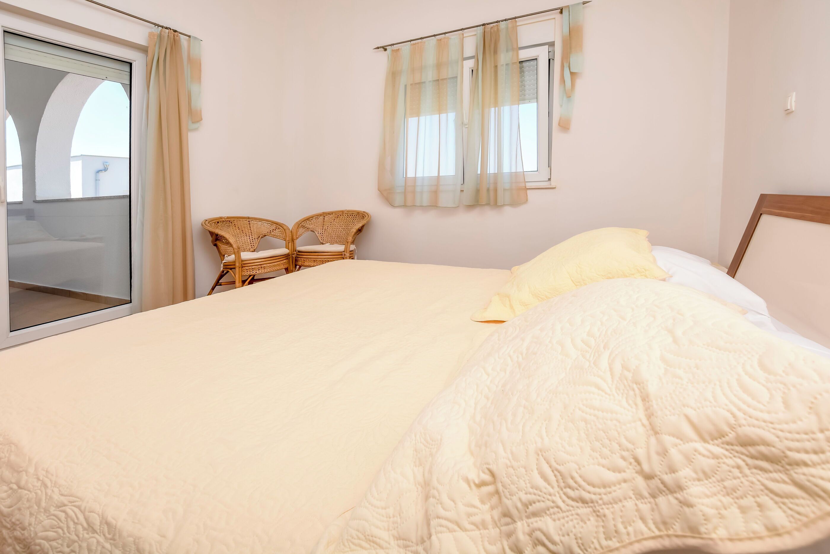 A bedroom with a large bed, a window with curtains, and two wicker chairs.