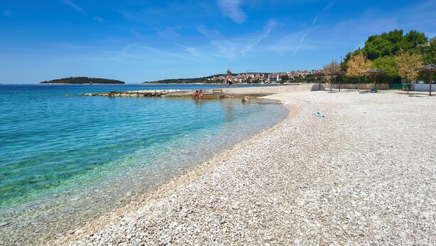 A pebble beach with clear water, a wooden swing, and a distant town. - Apartments Antea (Rogoznica)