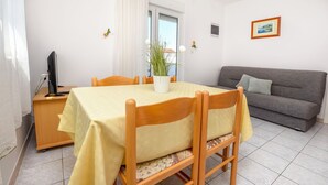 A dining area with a table set for six, a grey sofa, a TV, and a window with curtains. - Apartments Antea (Rogoznica)