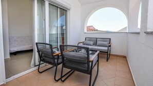 A balcony with two chairs and a table, a sliding glass door, and a view of a building with a red roof. - Apartments Antea (Rogoznica)