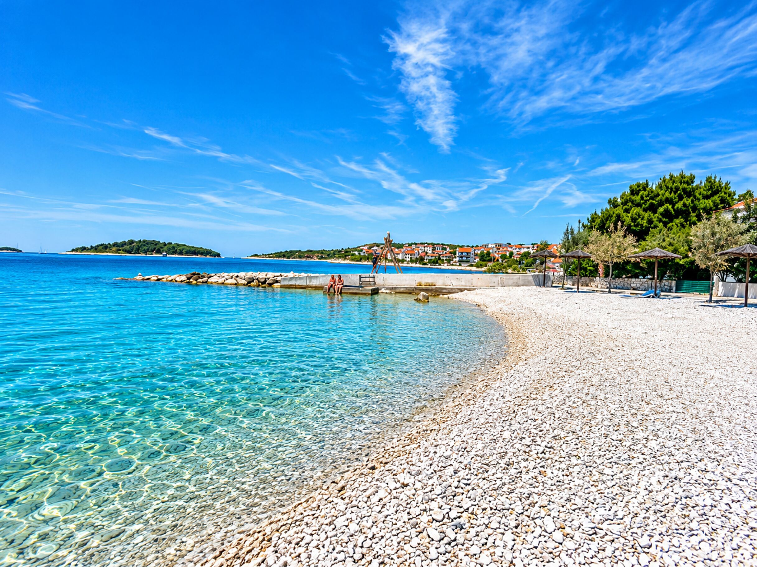 A pebble beach with clear water, a pier, and a village in the background.