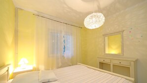 Un dormitorio con cama, mesita de noche con lámpara, cómoda, espejo y ventana con cortinas traslúcidas.
