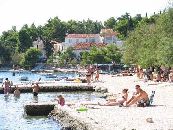 Ein Strand mit badenden und entspannenden Menschen, ein Poolbereich und Gebäude im Hintergrund.