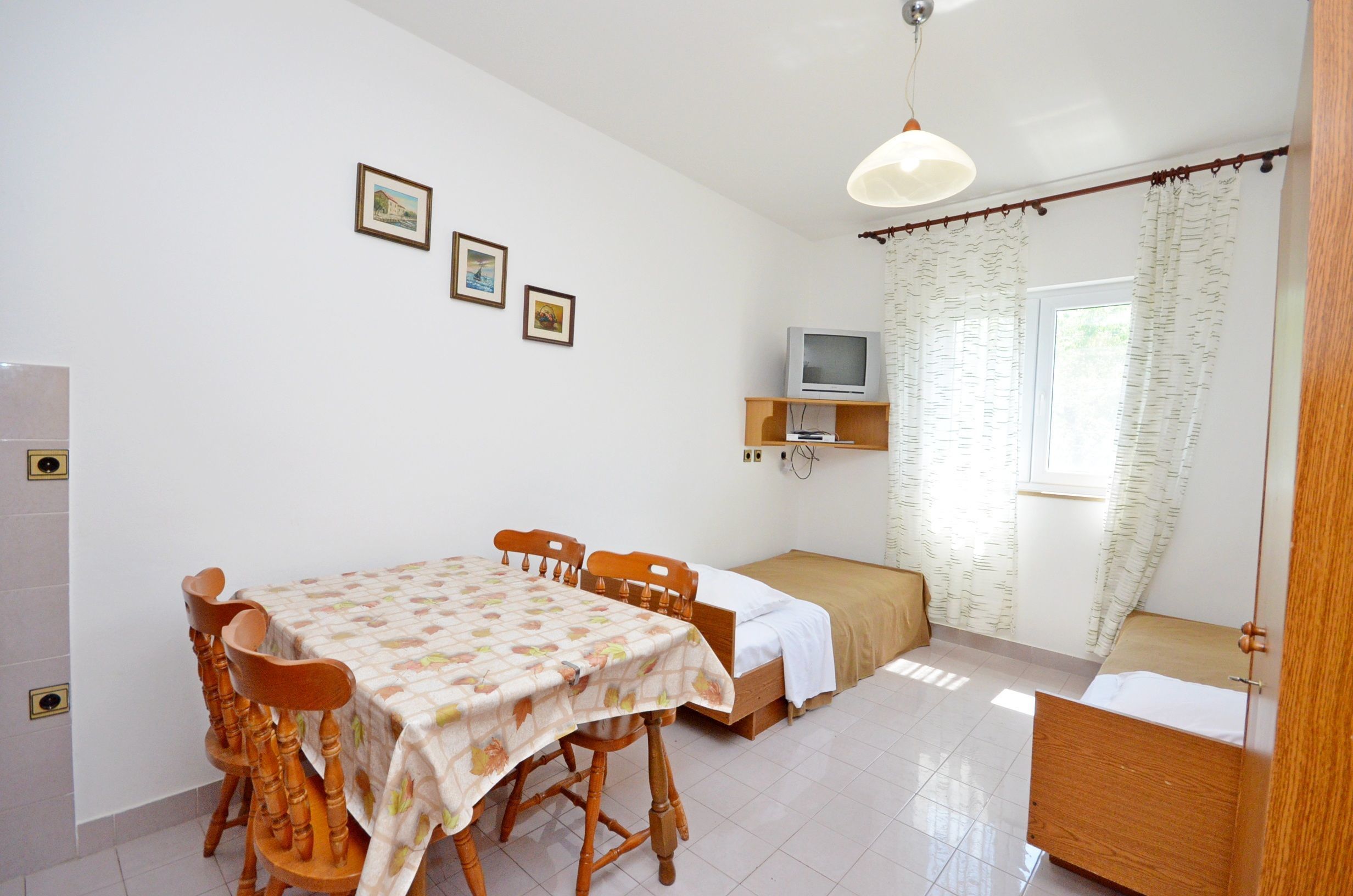 Una habitación pequeña y limpia con una mesa de comedor, sillas, una cama, un televisor y un ventanal con cortinas.