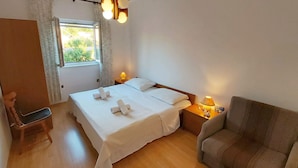 A neatly made bed with white linens and towels, a wooden chair, a sofa, a nightstand with a lamp, and a window with curtains. - Apartments Vila Punta (Jadrtovac)