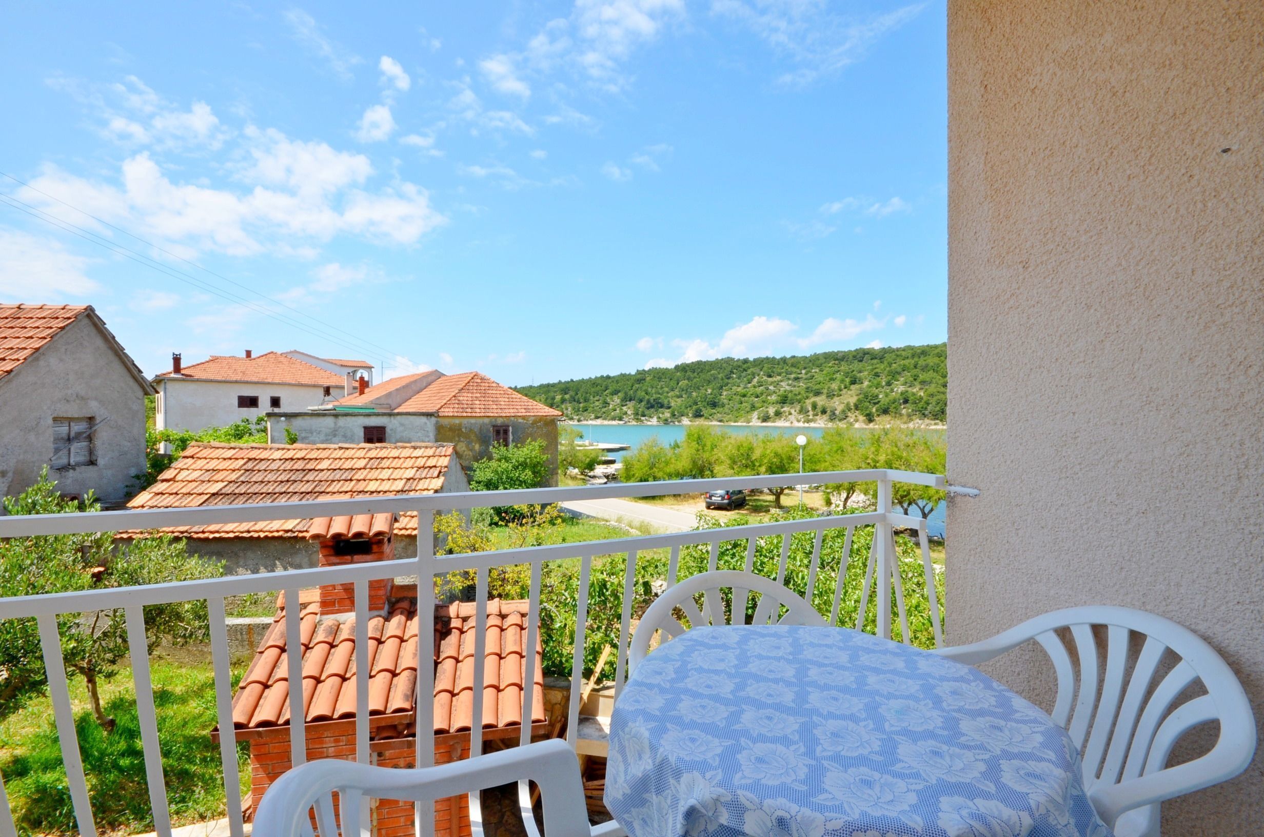A balcony with a table and chairs, overlooking a village with houses and a body of water.