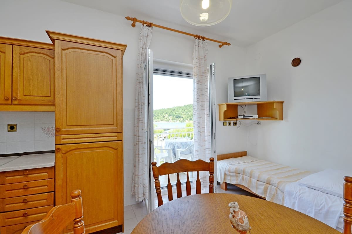 A room with a wooden dining table, a single bed, a TV on a shelf, and a window with curtains.