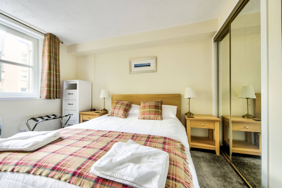 A neatly made bed with a plaid blanket, two bedside lamps, a small bedside table, a mirror, and a window with curtains.