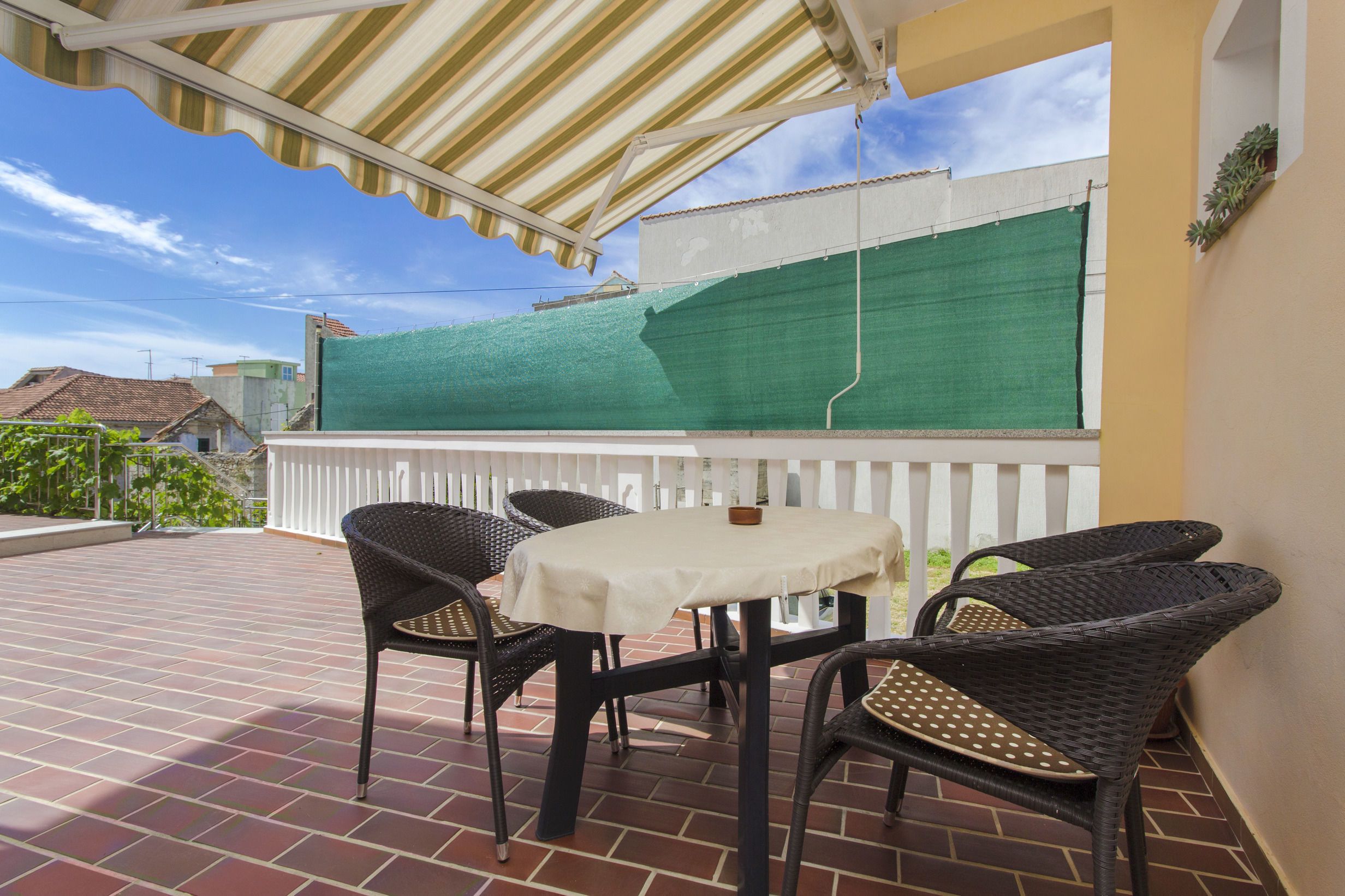 An outdoor patio with a table and four chairs, a green screen, and a white fence.