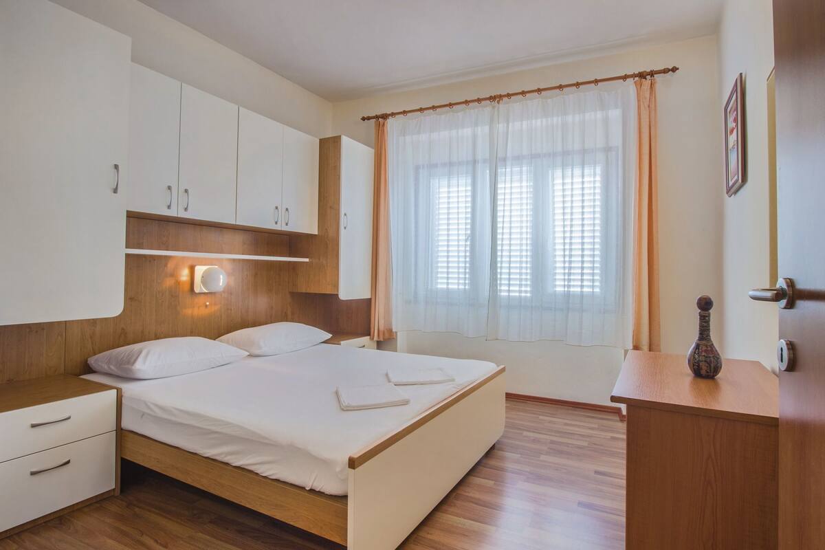 A bedroom with a wooden bed, white bedding, a nightstand, a dresser, and a window with curtains.