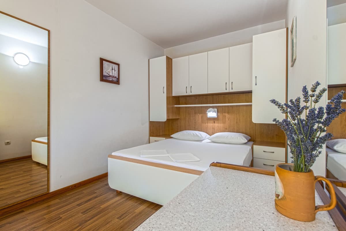 A bedroom with a wooden floor, white bed, and a nightstand with a vase of flowers.