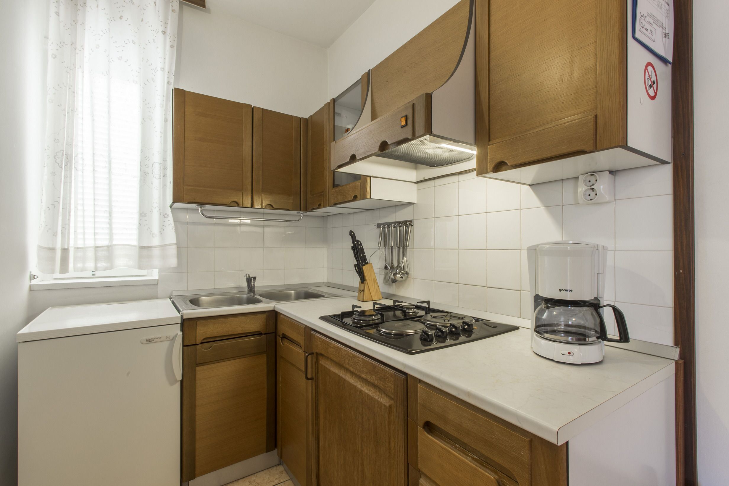 A compact kitchen with wooden cabinets, a gas stove, a coffee maker, and a window with a curtain.