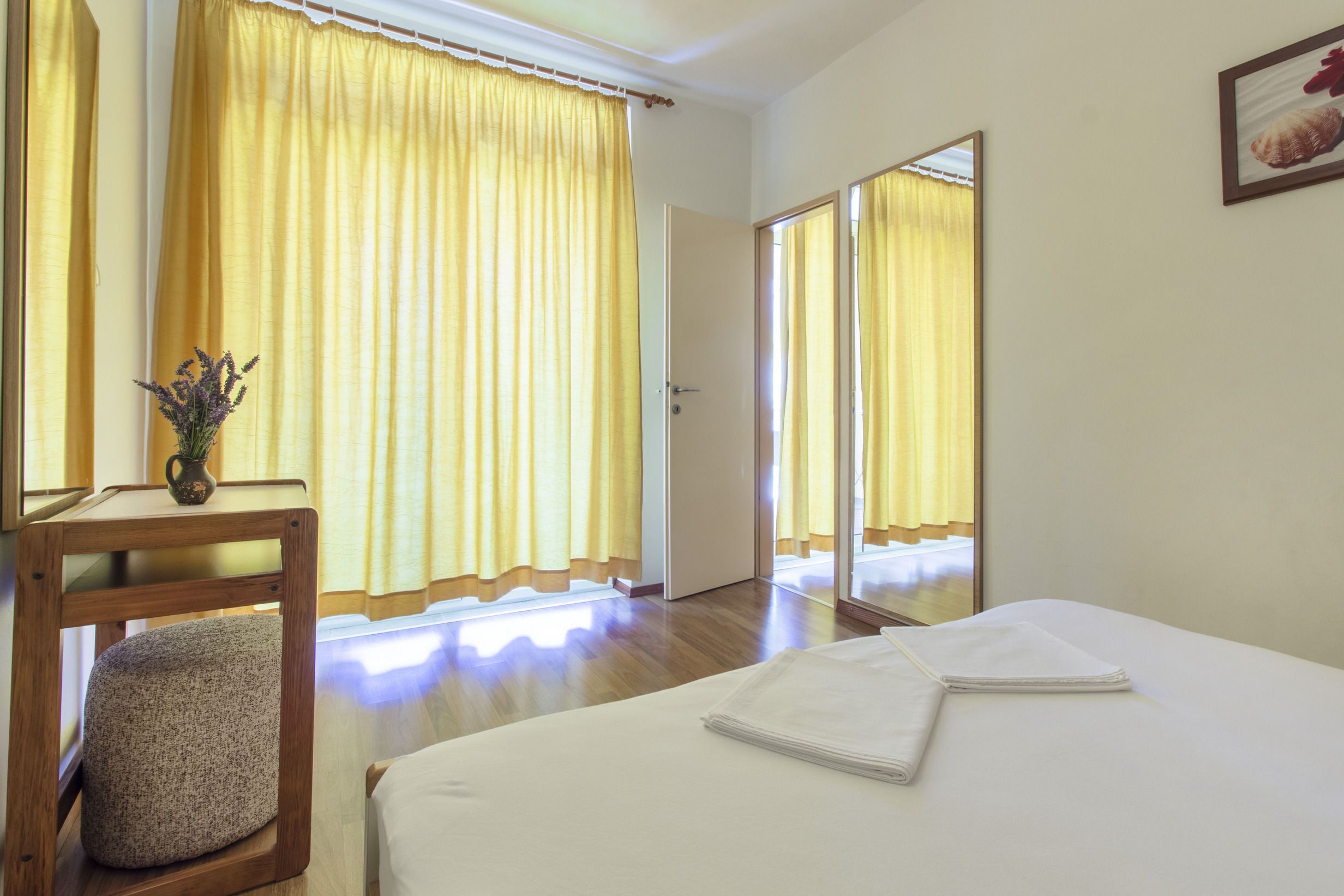 A hotel room with a bed, a wooden chair, a small table with a vase, and a mirror on the wall.
