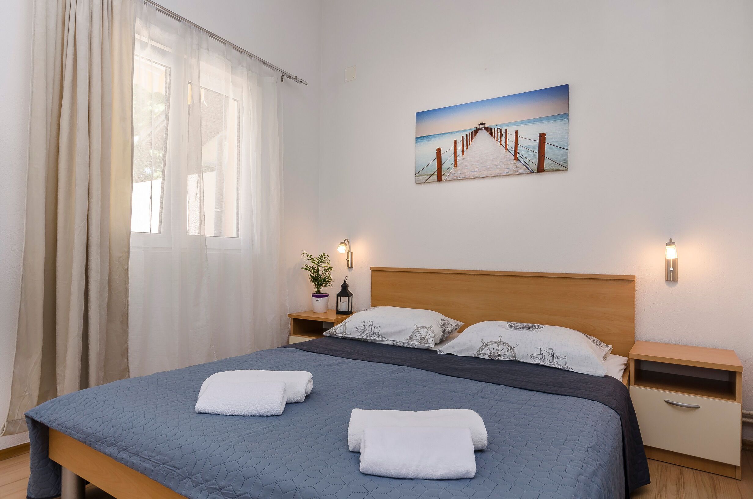 A neatly made bed with a blue quilt and white towels, a wooden headboard, a nightstand with a lamp, a wall-mounted painting of a pier, and sheer curtains on a window.