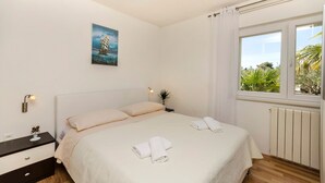 Una cama bien hecha con ropa blanca y toallas, una mesita de noche con una lámpara, una planta pequeña y un cuadro de un barco en la pared.