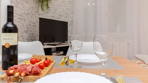 Una mesa con un servicio para comer, una botella de vino Plavačhvar, una tabla de madera con embutidos y tomates, y dos copas de vino.