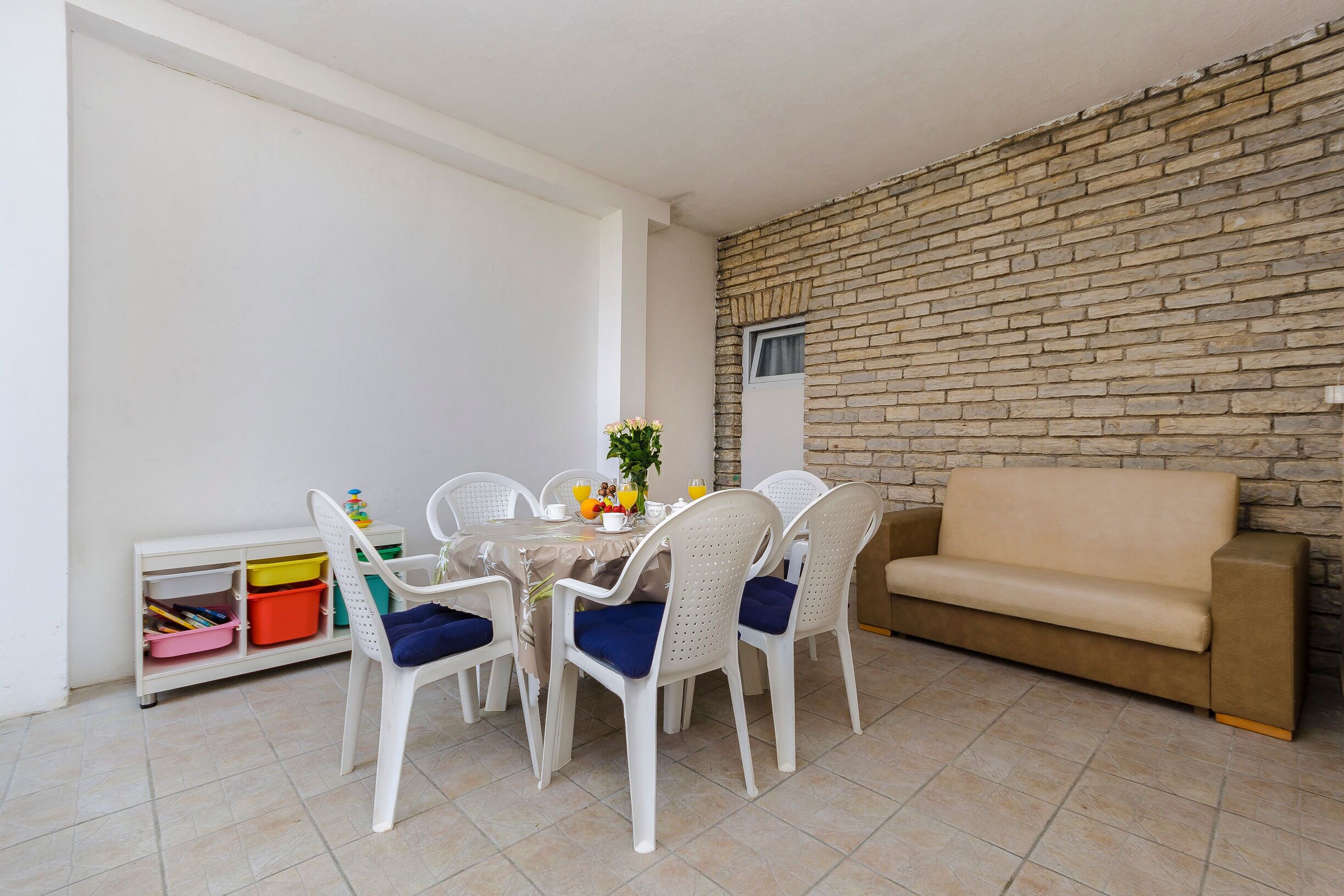 A dining area with a table set for four, surrounded by white chairs with blue cushions, a beige sofa, and a brick wall.