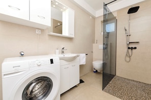 A modern bathroom with a washing machine, sink, toilet, and glass-enclosed shower. - Apartments Ante (Vodice)