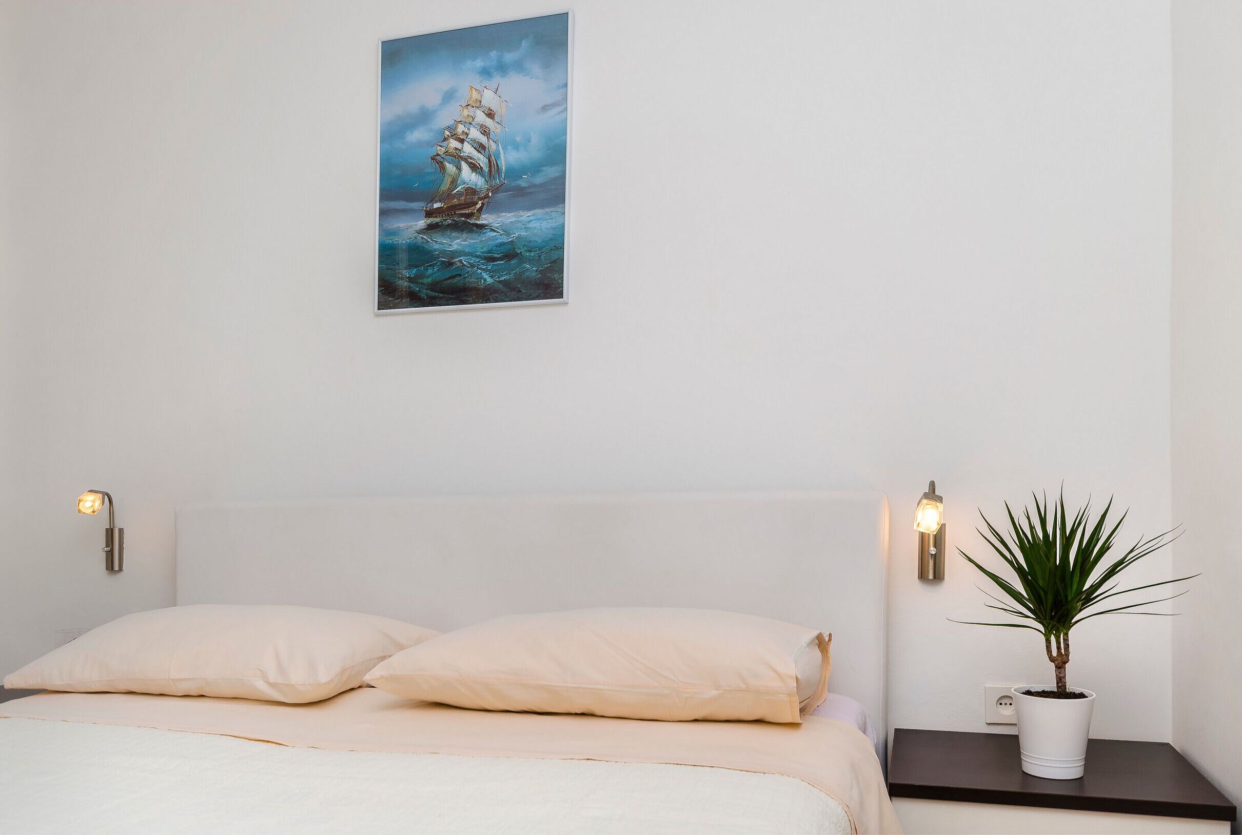 A bedroom with a bed, a nightstand with a plant, and a painting on the wall.