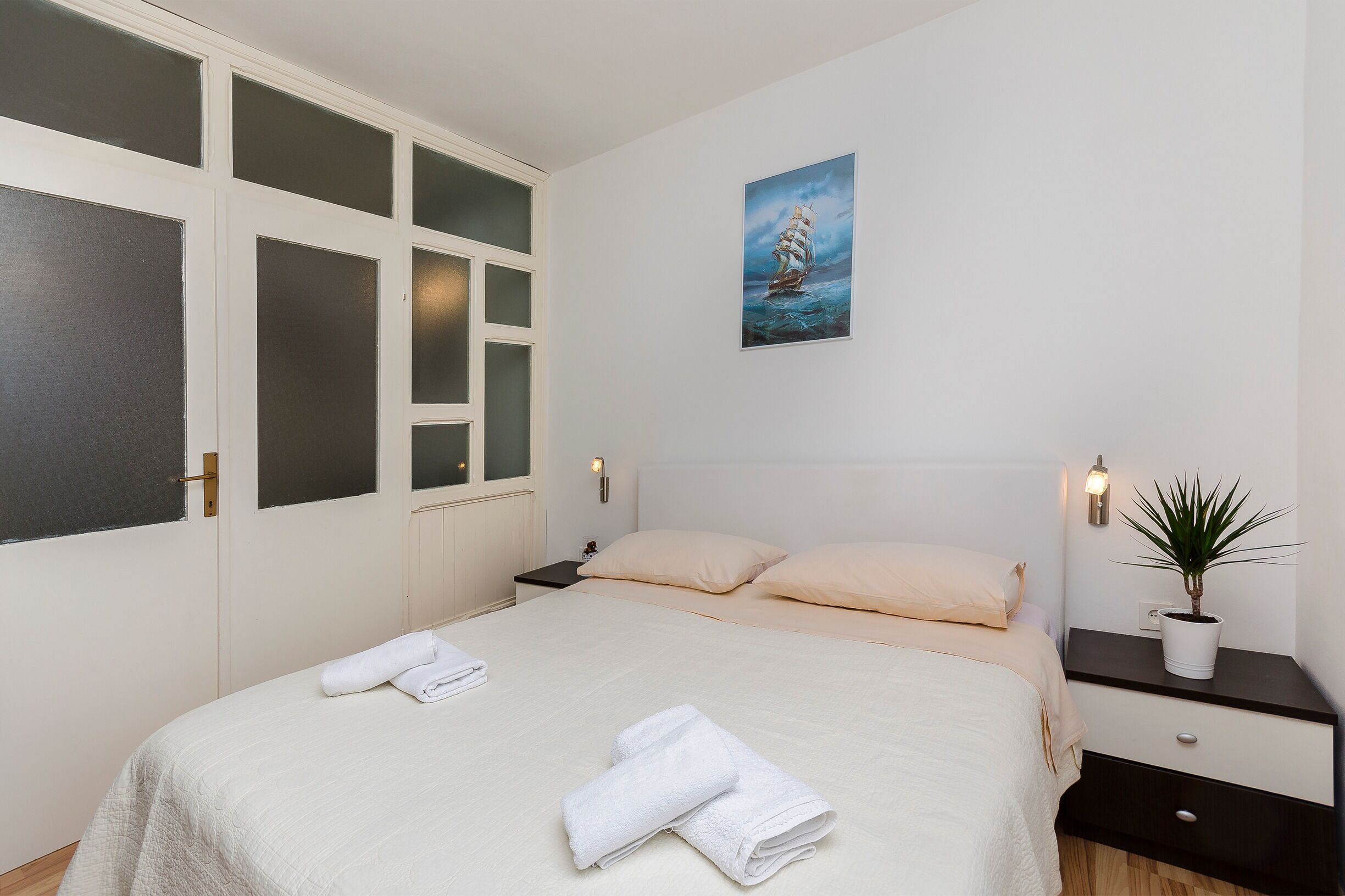 Un dormitorio con una cama, una mesita de noche con una planta, un cuadro en la pared y un ventanal con paneles traslúcidos.