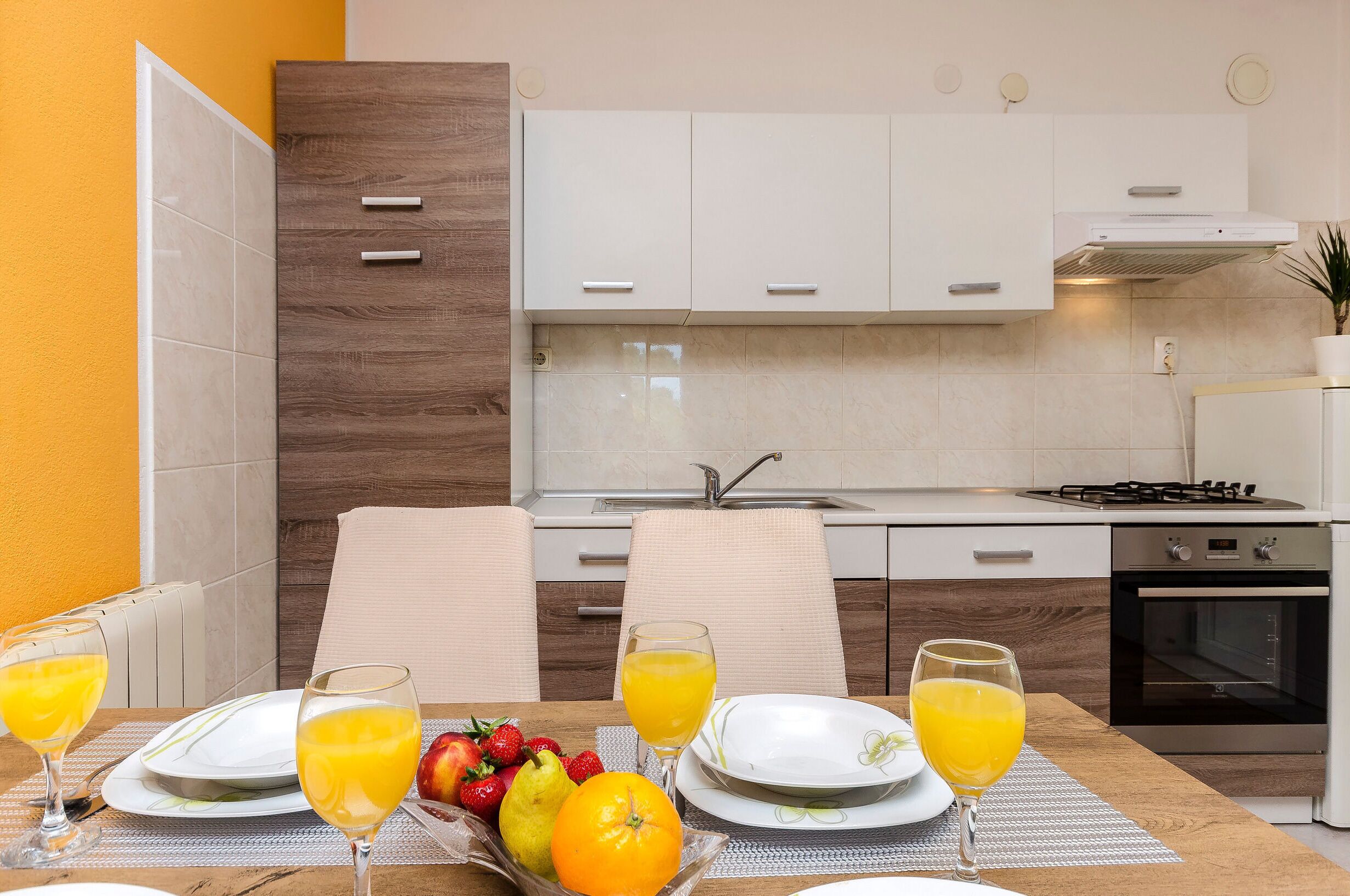 A modern kitchen with white cabinets, a gas stove, and an oven. There is a dining table with plates, glasses, and fruit.