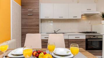 A modern kitchen with white cabinets, a gas stove, and an oven. There is a dining table with plates, glasses, and fruit.