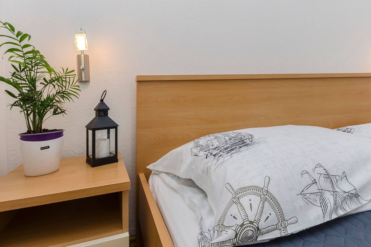 A neatly made bed with a patterned blanket, a bedside table with a potted plant and a lantern, and a wall-mounted light fixture.