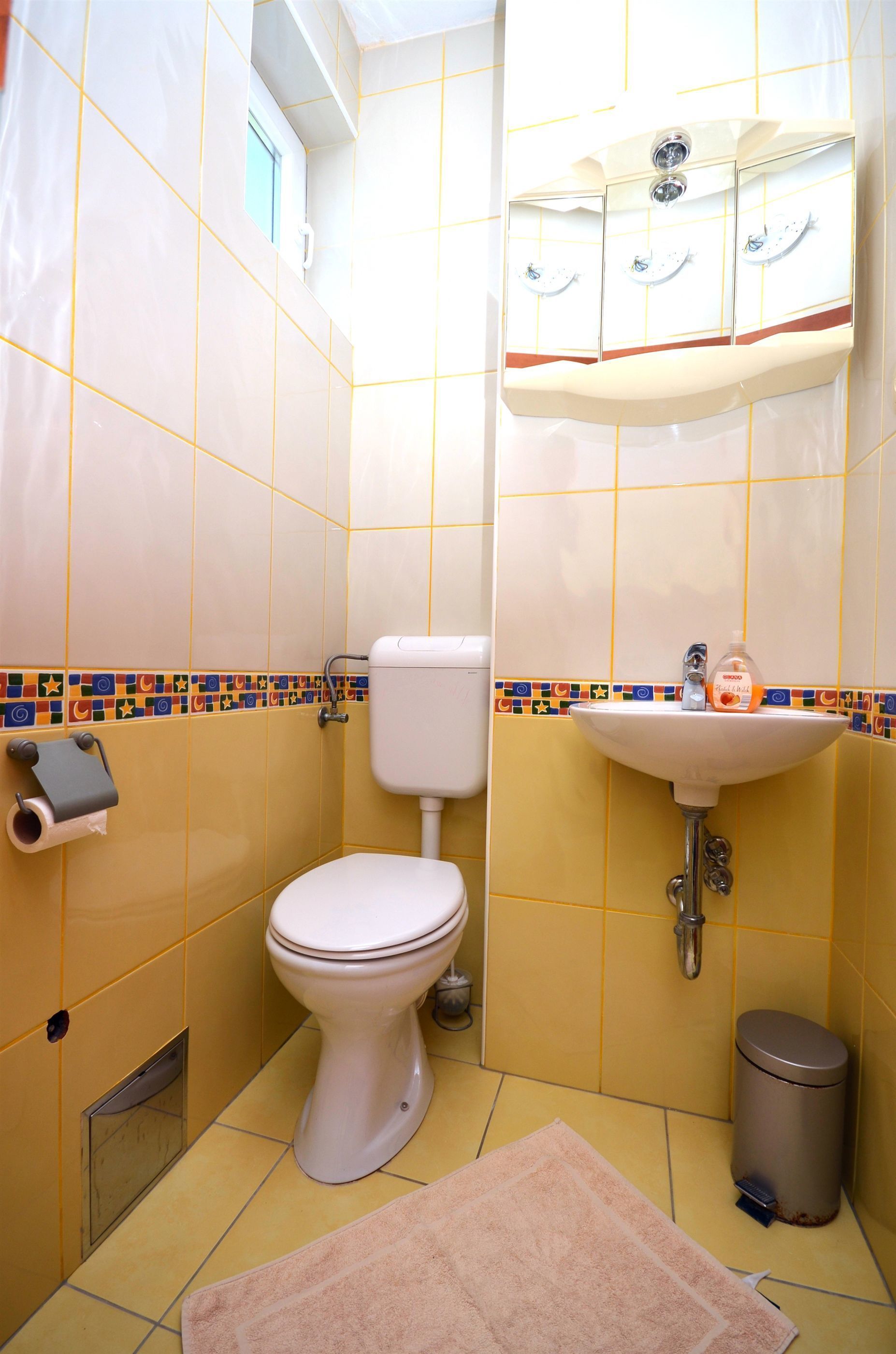 Un baño con azulejos amarillos, un inodoro, un lavabo con jabón y un cubo de basura.