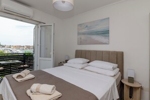 Et hotelværelse med en seng, et natbord, et lille bord, et maleri og udsigt over byen gennem en balkondør.