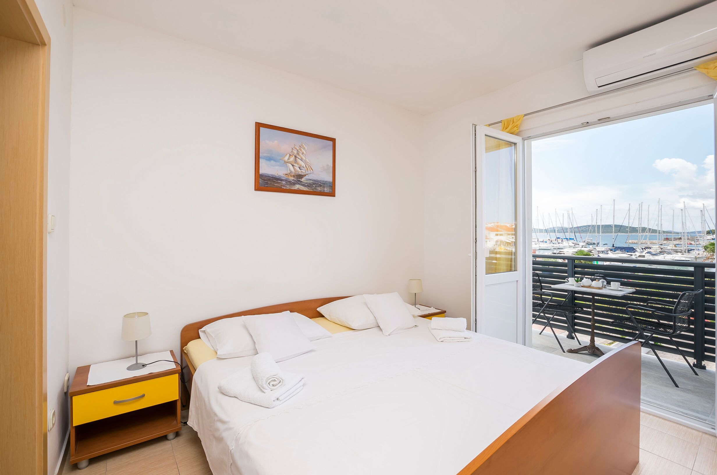 A hotel room with a bed, a nightstand, a window with a view of boats, and a painting on the wall.