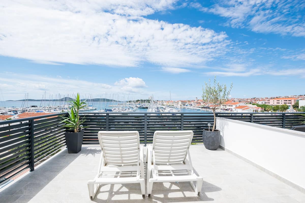 Um terraço com duas cadeiras de estar brancas, um corrimão e vista para uma marina com barcos e edifícios ao fundo.