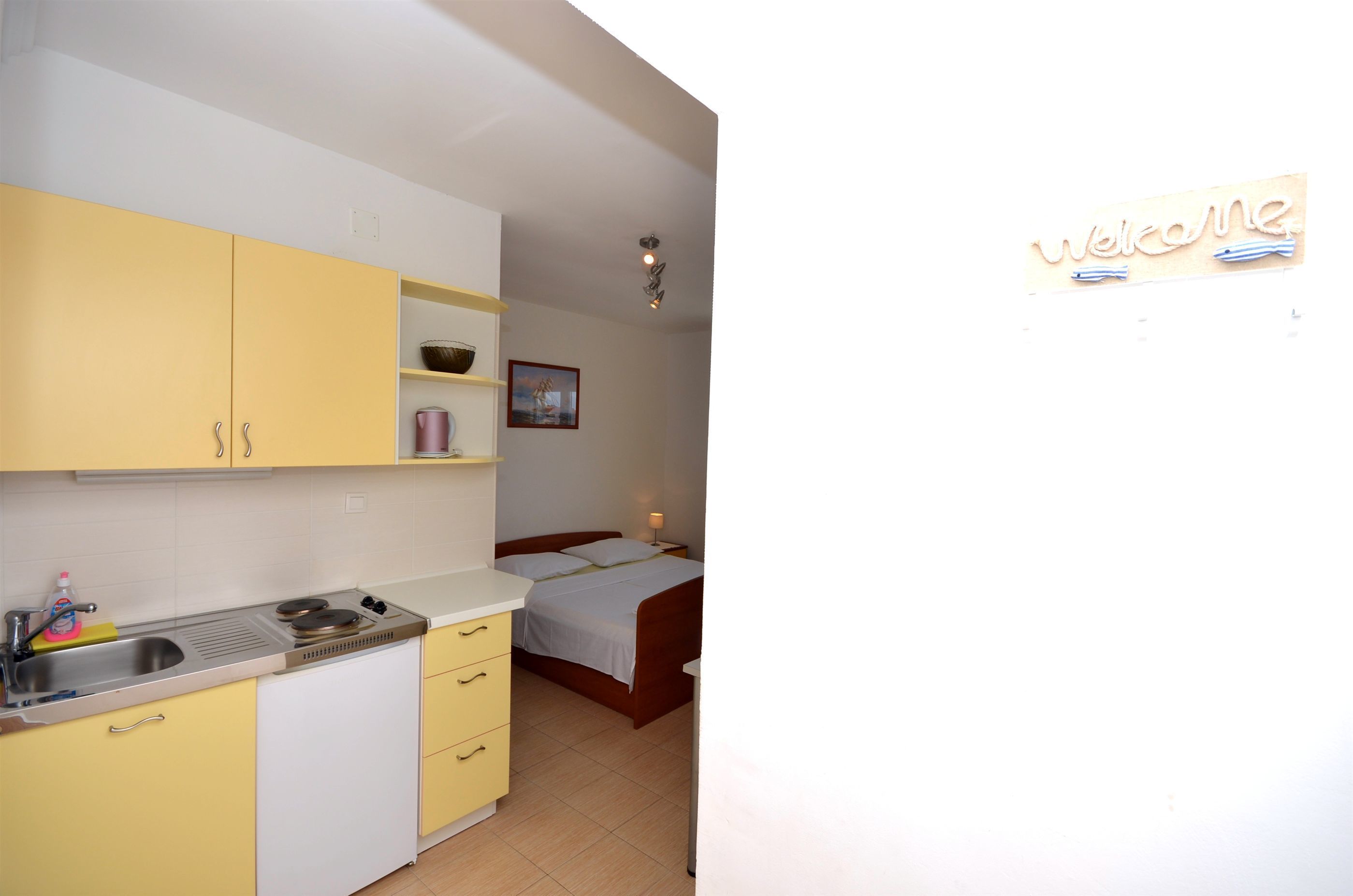 A compact kitchen with a sink, stove, and yellow cabinets. A bed with a white mattress and pillows is visible in the adjacent room.