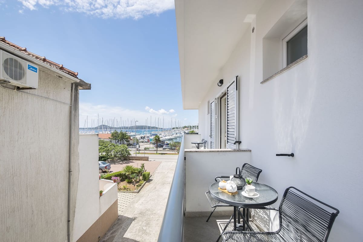 Uma varanda com mesa e cadeiras, com vista para uma marina com barcos e uma paisagem acidentada.