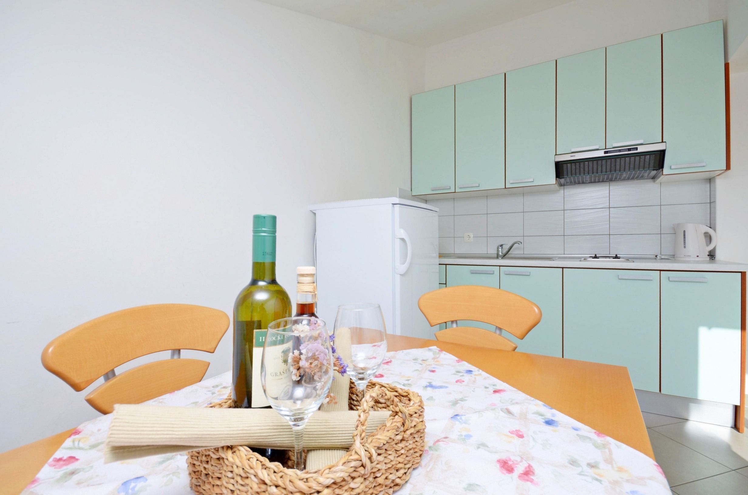 Une cuisine avec des meubles de couleur vert clair, un réfrigérateur blanc et une table dressée avec une bouteille de vin, des verres et un panier en osier.