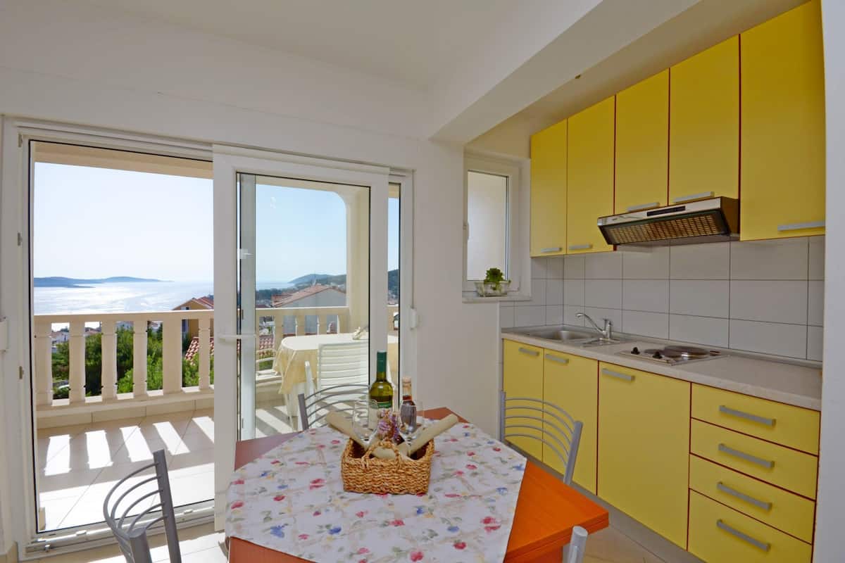 Un balcón con mesa lista para comer, una cocina con armarios amarillos y vistas al mar.